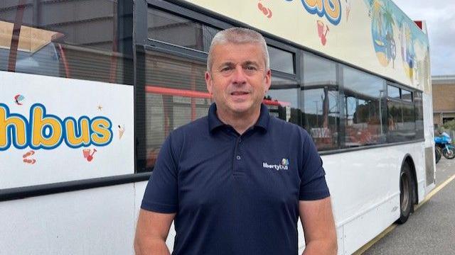 Kevin Hart who is wearing a blue shirt with the Liberty Bus logo and standing in front of one of the firm's liveried buses