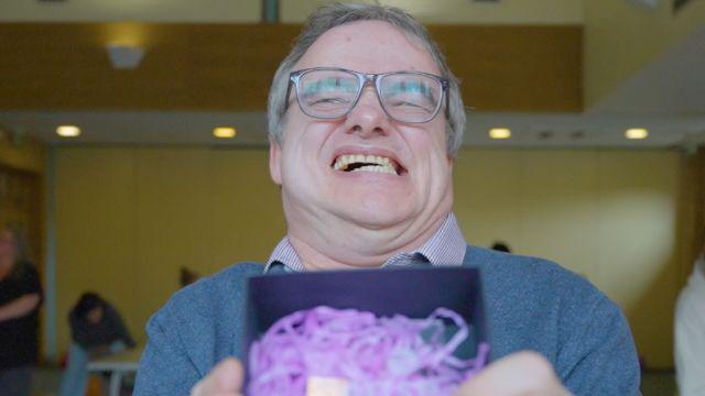 A man smiling broadly, baring his teeth. He is holding a trophy inside a box with purple shredded tissue. He is wearing a blue jumper and a checked shirt.