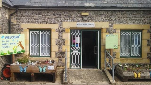 External view of the Merle Boddy Centre in Swaffham. The flint building has a ramp with grab rails leading to the green door. There are planters decorated with colourful butterflies outside.
