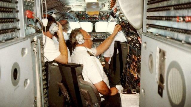 Inside a plane's cockpit with aircraft staff flicking buttons in a narrow corridor.