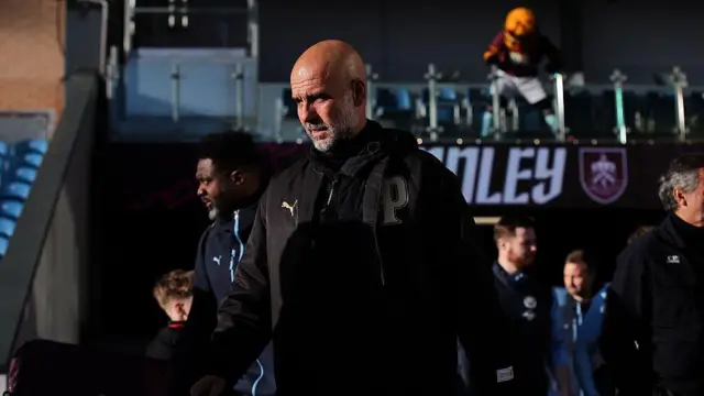 Pep Guardiola dressed in all black coming out of the players tunnel at Burnley, the sun shines on him.