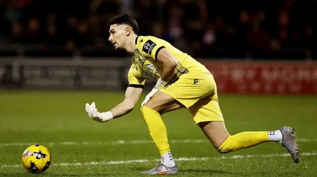 Luke Southwood, wearing an all-yellow kit, bowls the ball out along the ground for Bristol Rovers