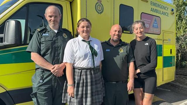 Two men in dark green shirts with NHS and South East Coast Ambulance Service logos on them standing next to a woman with long, tied-up hair, a black jumper and black bicycle shorts, and a teenage girl in a grey chequered skirt, white shirt and green and black tie. They are standing in front of an ambulance.
