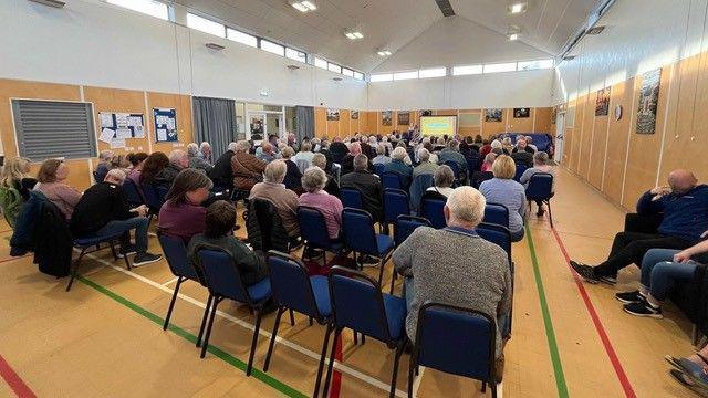 Rows of chairs with about hundred people in a village hall 