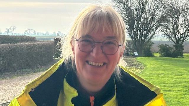 Joanna Sidgwick, 57, has blonde shoulder length hair with a bob over her forehead she is wearing spectacles and a high visibility yellow jacket with a dark fleece top underneath, behind her is farmland and trees.