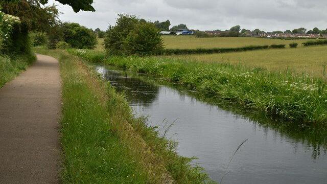 A path runs alongside a river that has formed in the remnants of the Manchester, Bolton & Bury canal. A bird can be seen wading in the river, which is flanked on the opposite side with green fields, with houses seen in the far distance.