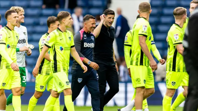Portsmouth manager John Mousinho walks on the pitch and talks with his players and fellow coach. The players wear a luminous yellow kit and Mousinho wear a black quarter zip top with black trousers
