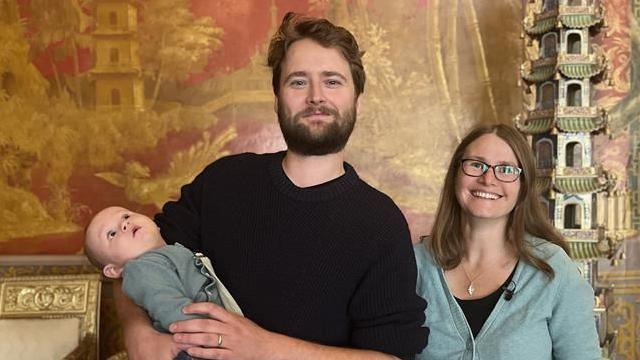 A bearded man dressed all in black is standing in front of a lavish red and gold Chinese patterned wallpaper, holding his baby in his arms. The baby is looking up at the ceiling. The man is looking into the camera, alongside a smiling woman wearing a black dress and a light blue cardigan. There is an ornamental pagoda in the background.