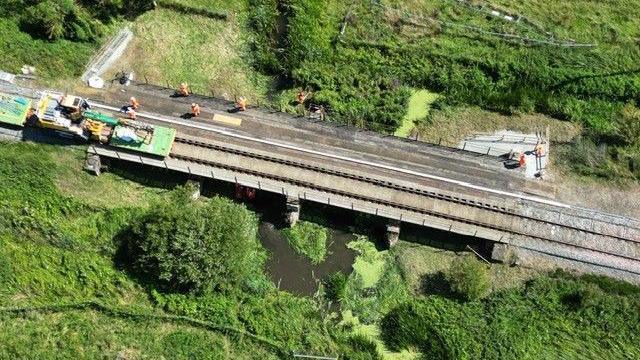 A railway going over a small river running through a field. There are people standing on the bridge and there is a construction vehicle on the left.