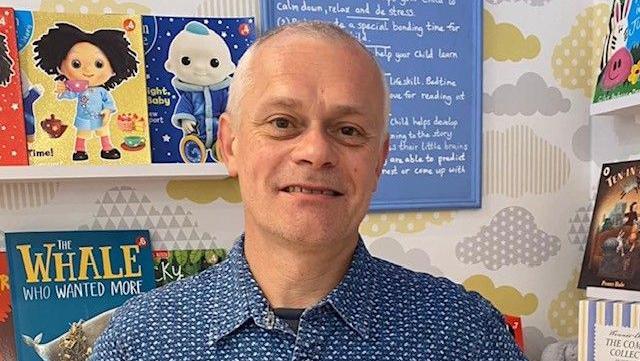Duncan Cooper has short grey hair. He is wearing a blue and white patterned shirt with a black T-shirt underneath. He is stood in his bookshop with children's books on white shelves behind him.