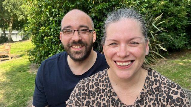 A close-up of Becky Anderson and her husband Matt in a garden with a lawn and large, green shrubs. Becky, who has swept-back, dark-grey hair, wears a leopard-print top and smiles as she looks into the camera. Matt, who is also smiling, has a shaven head, dark beard and wears dark-rimmed glasses and a blue T-shirt.