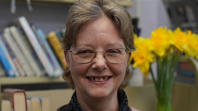 Rebecca Stephenson smiles at the camera. She has short blonde and grey hair and wears glasses. Behind her a base of daffodils can be seen as well as a bookshelf. 