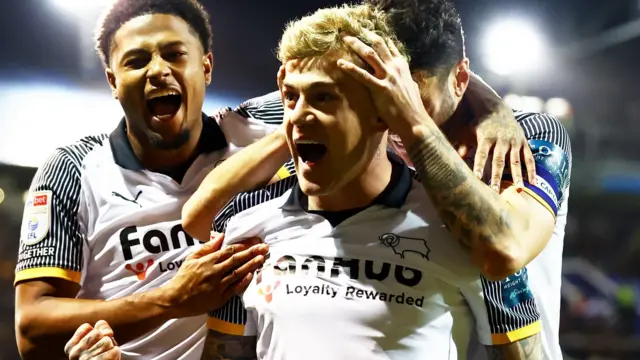 Sammie Szmodics punches his fist towards the fans after scoring the winner at Portsmouth, mouth wide open in celebration with one team-mate's hands on his head and another hugging him and shouting too