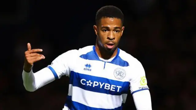 Elijah Dixon-Bonner on the pitch during a match in 2024 for QPR. He is wearing QPR blue and white striped kit and holding his right arm up with his palm pointing towards his body