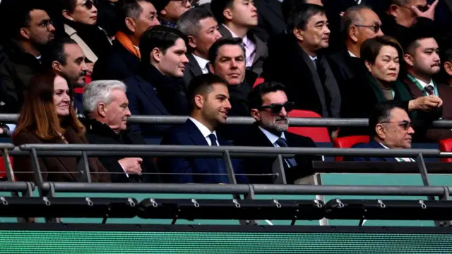 Jamie Reuben and Yasir Al-Rumayyan sitting in the stands surrounded by several other people