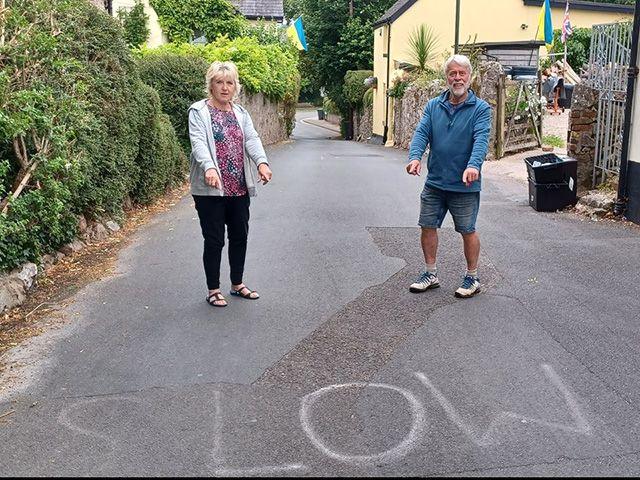 Marilyn and Norman Roberts stand on a narrow village road beside the word "SLOW" hand written on the tarmac, gesturing toward it. The street is lined with hedges and stone walls, with houses visible in the background.