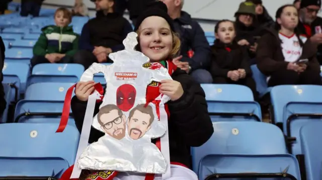 A young Wrexham fan holds up a foil FA Cup trophy with pictures of Ryan Reynolds and Rob McElhenney