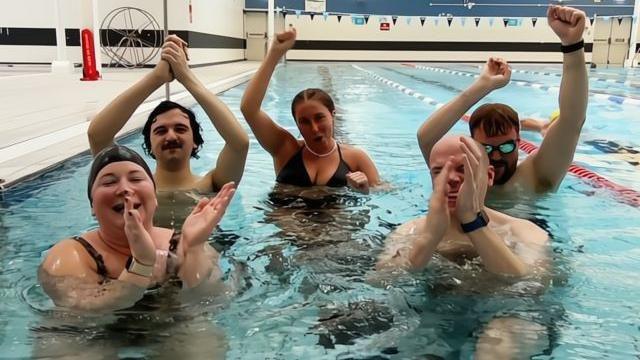 The five challengers are smiling and celebrating while submerged up to their shoulders in the swimming pool. Jordan wears reflective swimming goggles and his hands are triumphantly in the air.