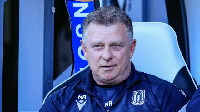 Stoke City boss Mark Robins watches from the dugout. He is wearing a dark blue club tracksuit.