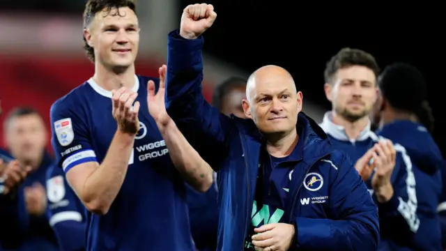 Alex Neil, wearing a blue Millwall jacket, raises his fist in the air to celebrate the win at Stoke in April, with captain Jake Cooper seen behind, in the navy blue Millwall home shirt, clapping his hands.