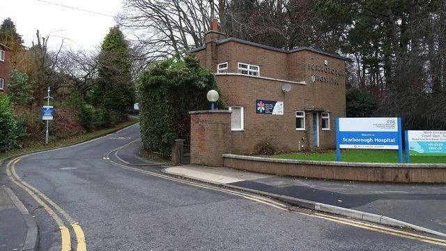 The North entrance to Scarborough Hospital, with a building reading "Scarborough Hospital", behind a blue and white sign with "Scarborough Hospital" written on it and an NHS logo.