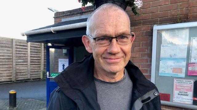 A photo of Jim Hardwicke outside the community centre. He is smiling to camera and wearing glasses. He is bald and is wearing a black coat with a grey t-shirt underneath.