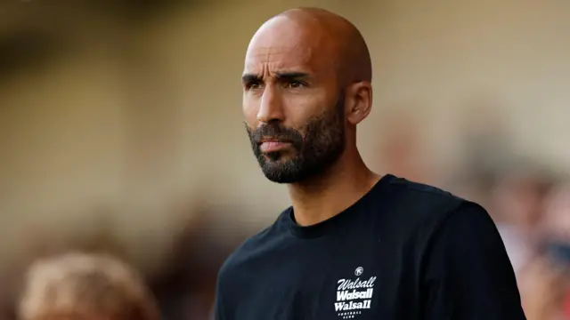 Walsall head coach Lee Grant watches his side from the touchline. He is wearing a dark sweatshirt with Walsall written in white on front.