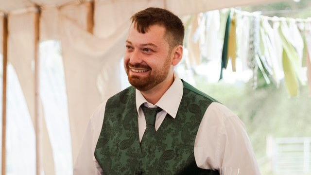 A man wearing a white shirt, green tie and green waistcoat smiling in a wedding tent.