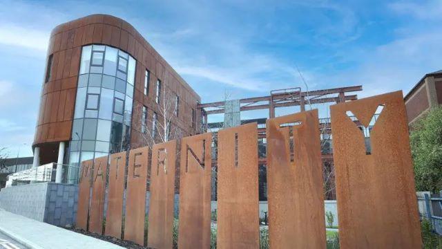 The maternity hospital at the Royal Victoria Hospital. ou can see a multi-story building with copper coloured cladding and large windows. In the foreground there is grey pavement and a copper coloured sign spelling out the word maternity. 