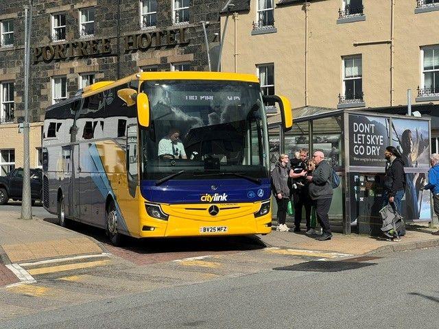 Bus buidhe agus gorm le daoine a' feitheamh airson a dhol air bòrd.