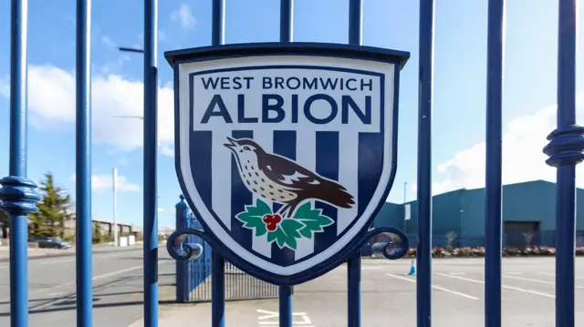 A photo of the West Bromwich Albion club crest on a blue gate at the Hawthorns with a parking lot and a road with houses in the background
