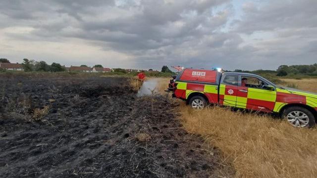 An all terrain fire brigade vehicle parked by a burnt area of grassland. A firefighter is hosing down undamaged grassland adjacent to the burnt area. 