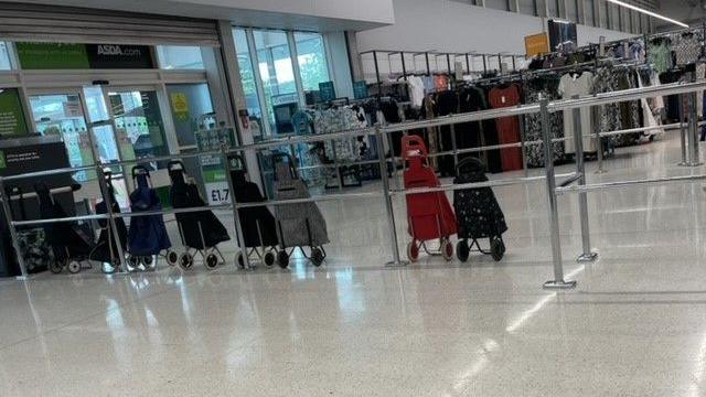 A row of shopping trolleys lined up next to railings inside an Asda supermarket, with dresses on railings in the background