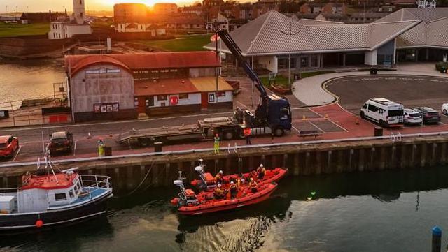 Two small lifeboats sit in a harbour. One is being lifted in by crane
