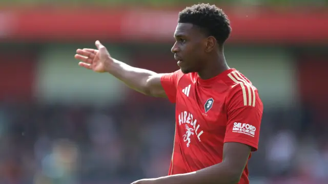 Salford City's Loick Ayina, wearing the club's red home shirt, orchestrates his defence with his right arm outstretched