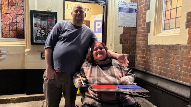 Emma and Wes are outside their local pub. Emma is in her electric wheelchair while Wes is standing next to her on the left of the picture.