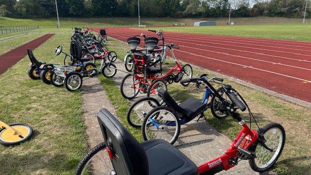 A row of trikes and bikes of various sorts lined up on grass alongside a red running track