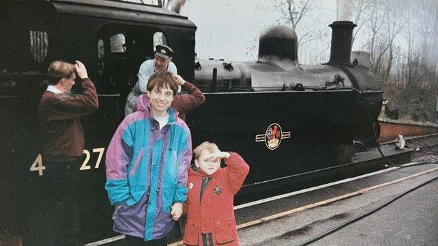 An photograph taken in the 1990s of Christine McCulloch wearing a raincoat and a young Adam wearing a red coat as the pair pose next to a steam train 