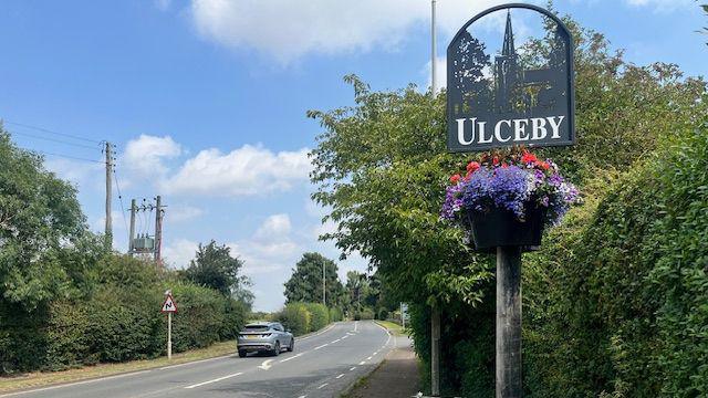 A road in Ulceby, North Lincolnshire. A silver car is travelling along the road which is lined by bright green hedges. There is a sign with the name of the village and brightly coloured flowers around it.