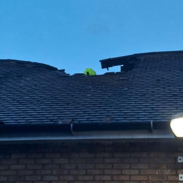 Damaged tiled roof with a large hole near the ridge, as a firefighter in high-visibility clothing inspects the hole.
