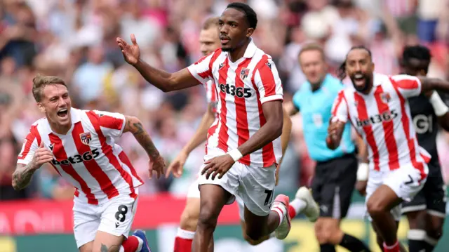 Eric Bocat, wearing Stoke City's red and white striped home kit, celebrates scoring against Swansea. His team-mates Ethan Galbraith, left, and Sorba Thomas, right, can be seen running either side of and just behind him.
