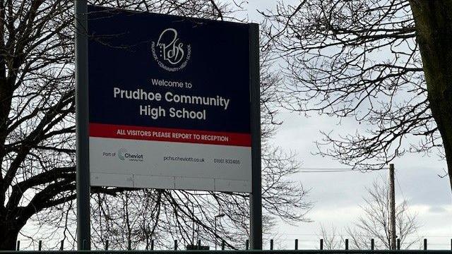 A sign saying Prudhoe Community High School is on two poles and dominates the picture. It is a cloudy day in winter and behind are trees without leaves 