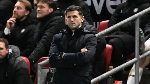 John Mousinho standing on the side line of the football pitch with his arms crossed, and looking disappointed.