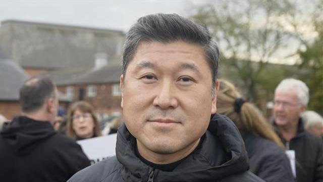 Gordon Young smiles at the camera. He has dark hair and wears a black puffer jacket. Protesters can be seen talking behind him.