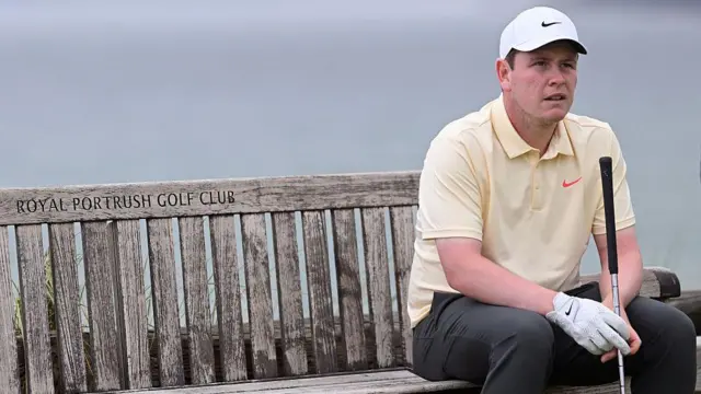 Scottish golfer Robert MacIntyre sits on the famous bench at Portrush. He is wearing a white cap and wearing a yellow t-shirt. He's sat on the right hand side of the screen and the left hand side shows the bench with the inscription 'ROYAL PORTRUSH GOLF CLUB'.
