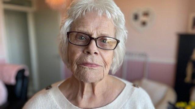 Mo Simmons looks directly at the camera. The background is slightly blurred behind her, but shows a pink living room. Mo has short grey hair and wears rectangular black framed glasses and a white round neck knitted jumper which has wooden buttons on the shoulders.