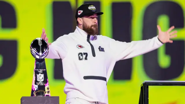 Travis Kelce plays up to the crowd in front of the Vince Lombardi Trophy at Super Bowl Opening Night media session