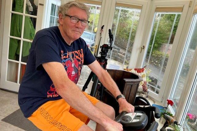 A man with grey hair and glasses sat in a conservatory in a dark blue shirt with bring yellow shorts. He is sat on an exercise bike which is slightly visible on camera