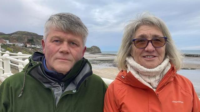 A man with blue eyes and grey hair in a green raincoat and a woman with grey hair and orange-tinted glasses in a thick white knitted jumper and red raincoat