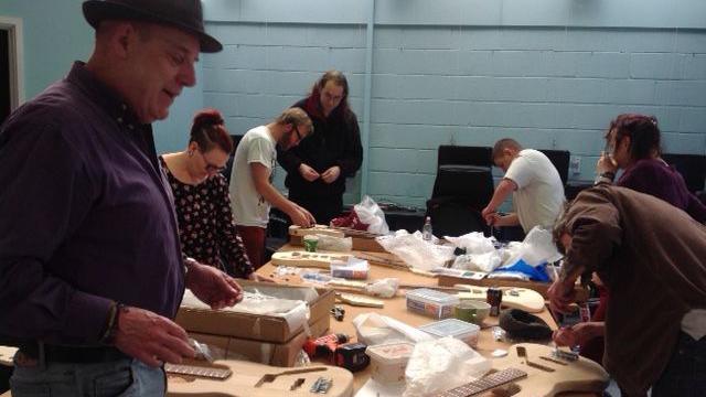 Seven people of different ages and genders gather around a woodworking table, building guitars.